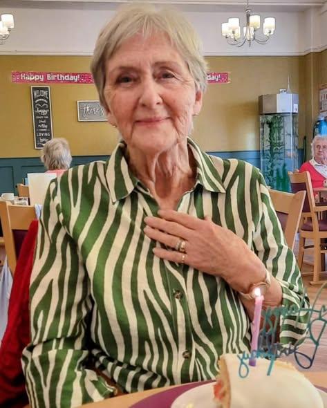 An elderly woman in a green and white striped shirt sits at a table with a pudding and lit birthday candle, in a decorated dining area.