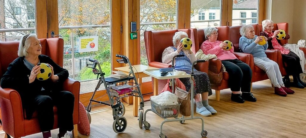 Five elderly women seated indoors on chairs, each holding a yellow and black ball. A walking frame and a trolley are nearby. Large windows reveal trees and buildings outside.