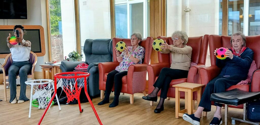 Four elderly individuals and an instructor sit on chairs, each holding a ball and aiming at toy basketball hoops in a bright indoor environment.