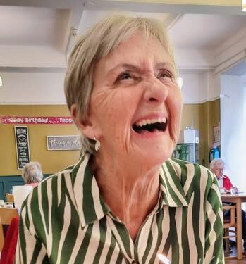 An older woman with short grey hair smiles at a table with a slice of cake and a lit birthday candle in front of her.