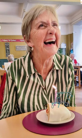 An older woman with short grey hair smiles at a table with a slice of cake and a lit birthday candle in front of her.
