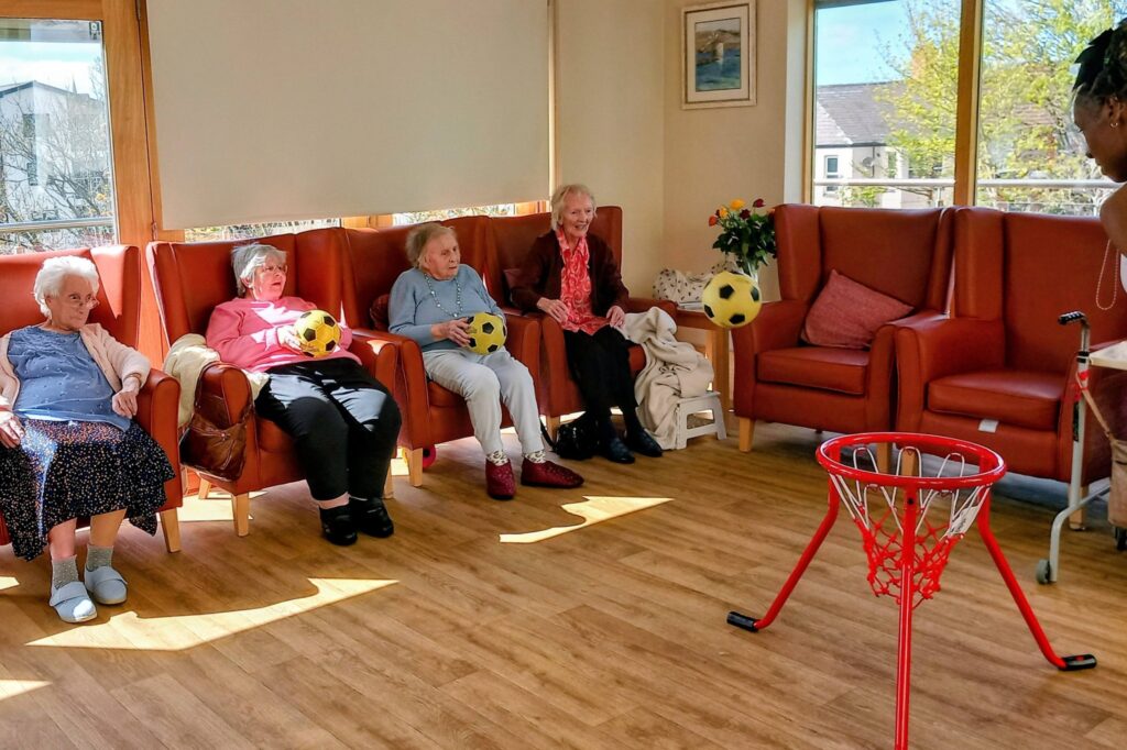 Four elderly women seated in armchairs each hold or toss a yellow football towards a small basketball hoop in a bright, wooden-floored common room.