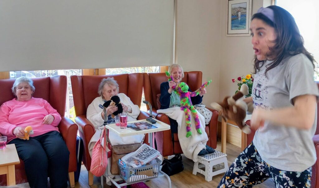 Three elderly women sit in armchairs holding cuddly toys, while a younger woman in pyjamas energetically engages with them in a brightly lit room.