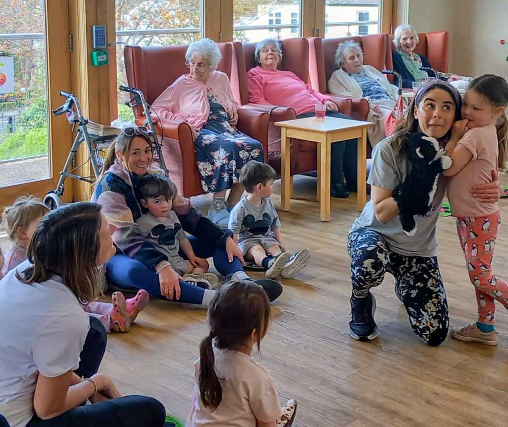 Young children and adults play together on the floor of a communal room while elderly women sit in armchairs, observing from the background.