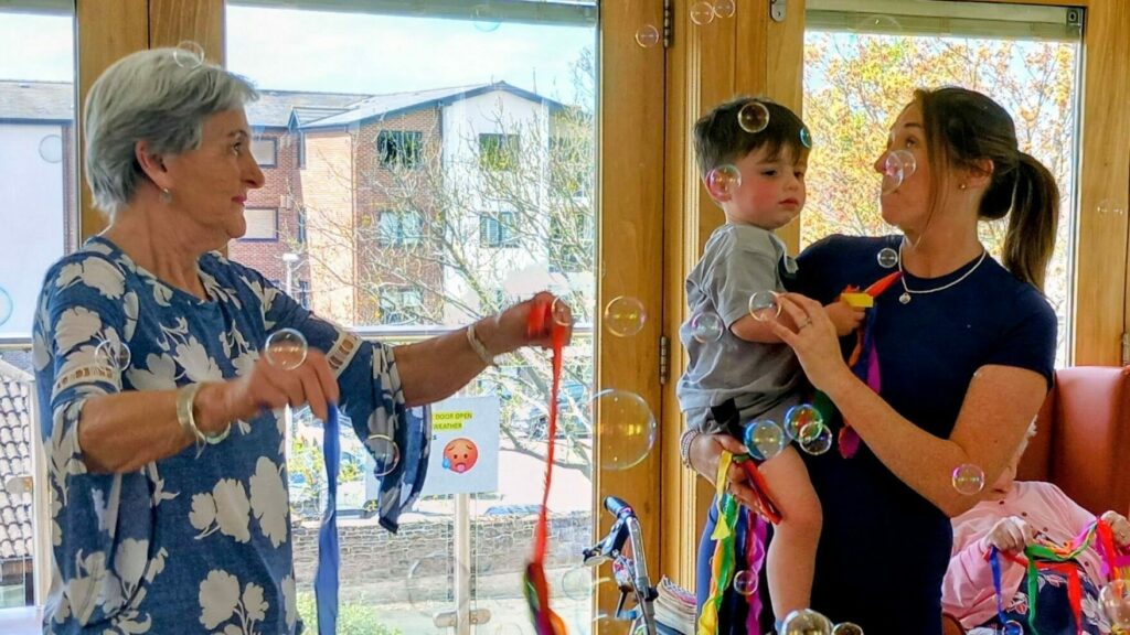 A woman holds a child as another woman waves colourful ribbons. Several children and soap bubbles fill the lively indoor setting.