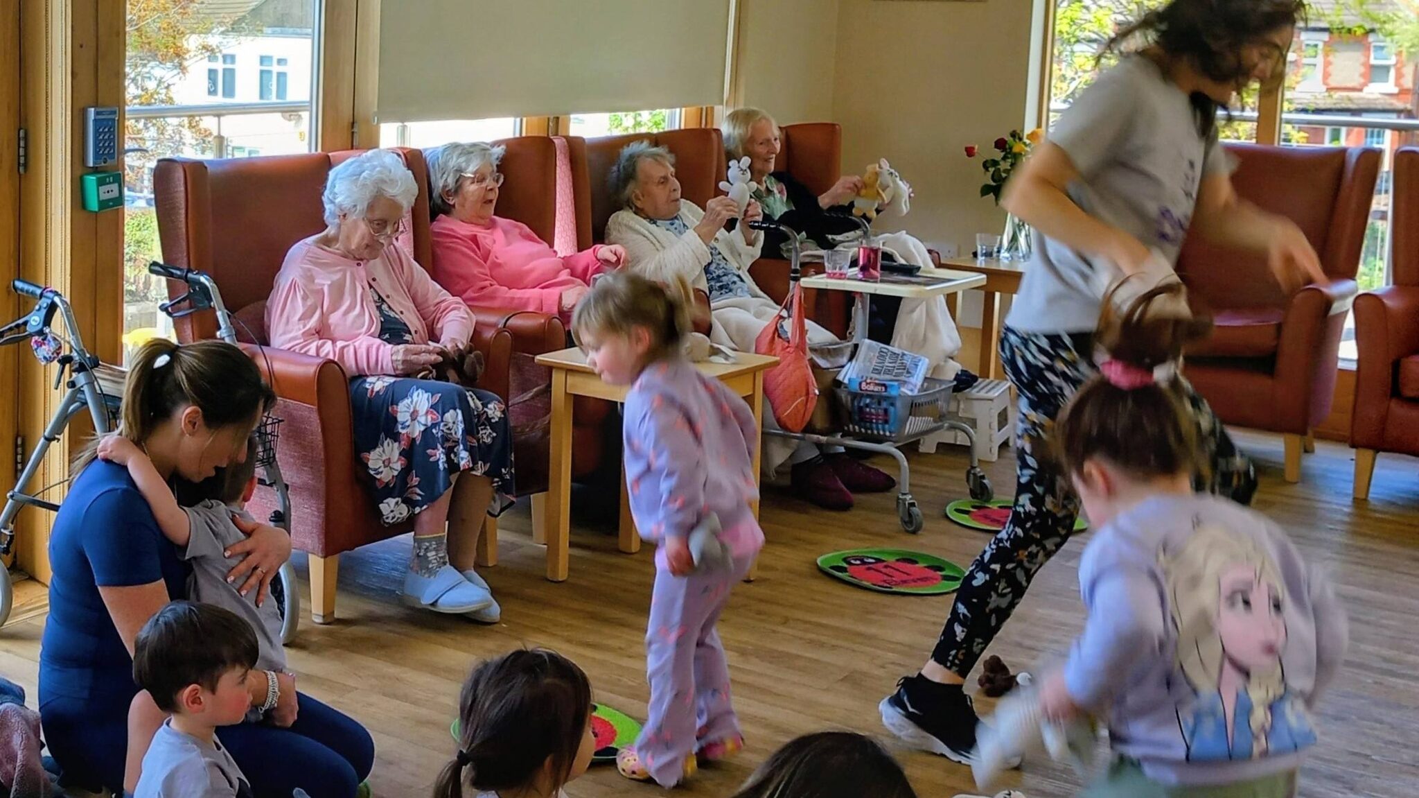 Children and elderly people engage with one another in a brightly lit communal room; some take part in activities while others observe from their chairs.