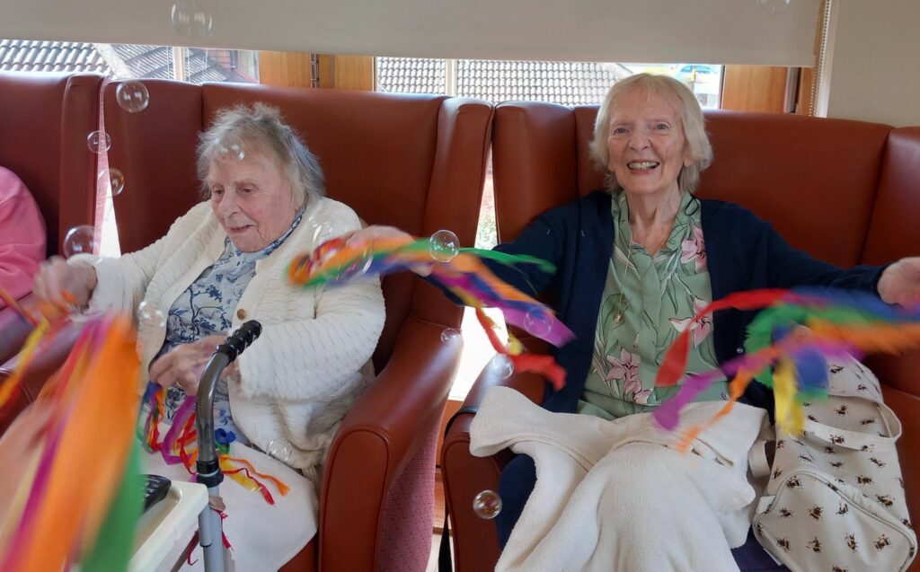 Two elderly women sit in armchairs, smiling and waving colourful ribbon wands, with soap bubbles floating nearby.