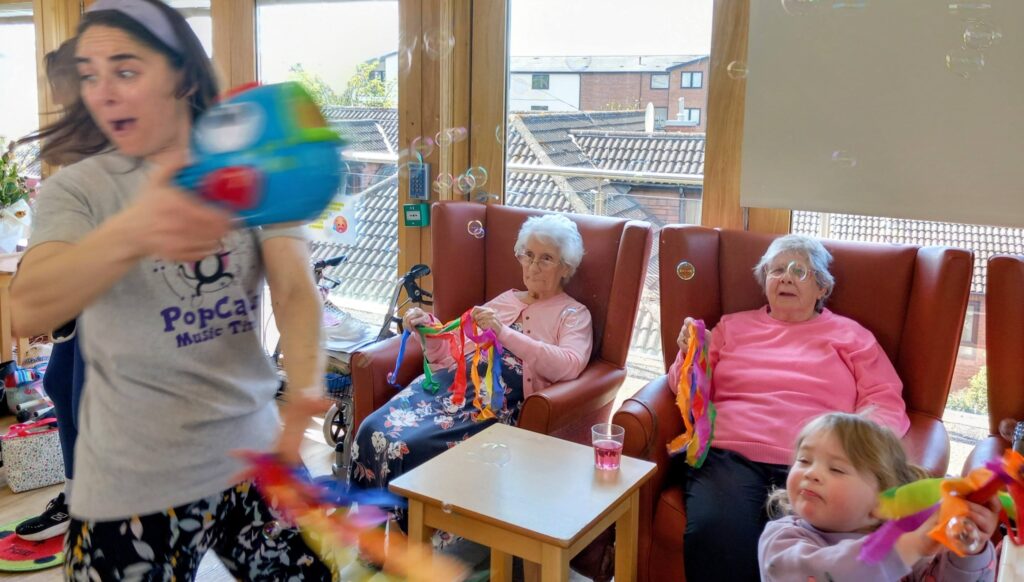 A woman waves a colourful streamer and operates a bubble machine near three older women and a child seated in armchairs, as bubbles float around the room.