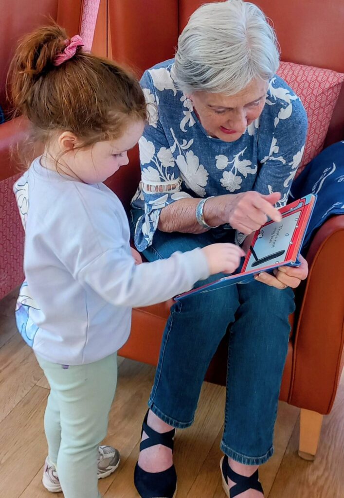 An elderly woman and a young girl sit together, engaging with a drawing pad as the woman sketches and the girl points at the screen.