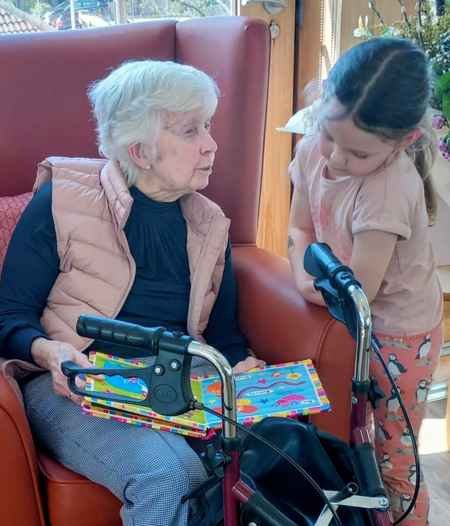 An elderly woman seated in a chair holds a colourful puzzle, whilst a young girl stands beside her, looking at the puzzle. A walking frame is positioned in front of them.