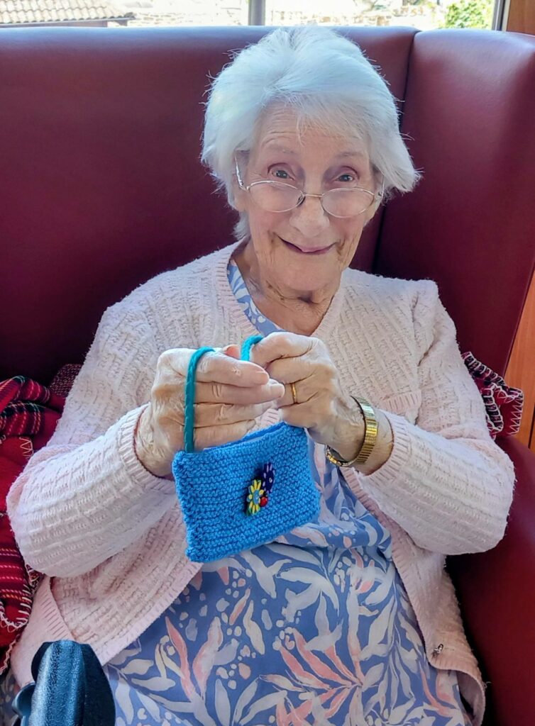 Elderly woman with glasses and white hair sits on a red chair, smiling and holding up a small blue hand-knitted purse adorned with a flower—one of many Shoebox Surprises that bring Stitches and Smiles.