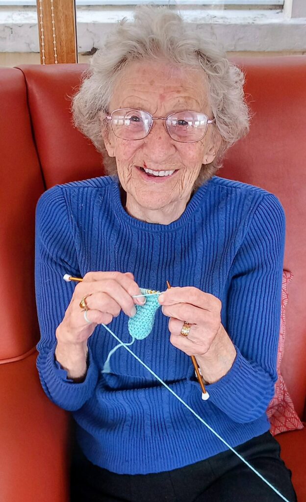 An elderly woman with spectacles, dressed in a blue jumper, sits on a red chair and knits light blue wool. Her stitches are neat and her smile is warm as she looks towards the camera.