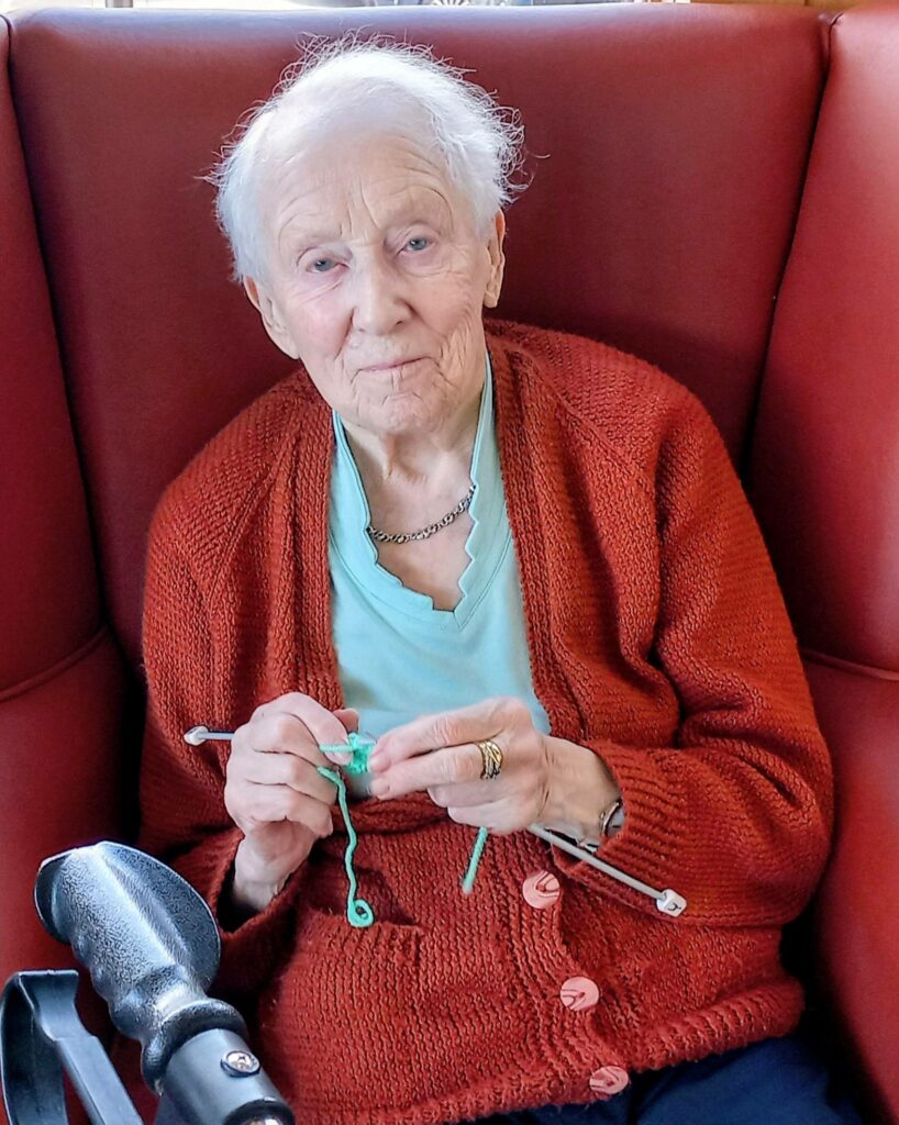An elderly woman in a red cardigan sits in a red chair, knitting green stitches with silver needles, her face aglow with warm smiles.