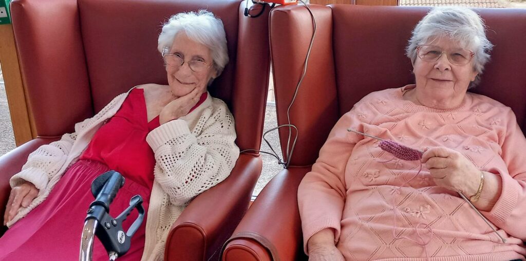Two elderly women sit in armchairs; one is dressed in a pink dress with a cream cardigan, whilst the other, holding knitting needles and pink wool, shares stitches and smiles during a cosy moment together.