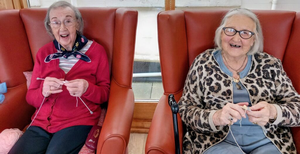 Two elderly women sit in red chairs, exchanging smiles and knitting together; their hands occupied with stitches and wool, crafting heartfelt Shoebox Surprises.