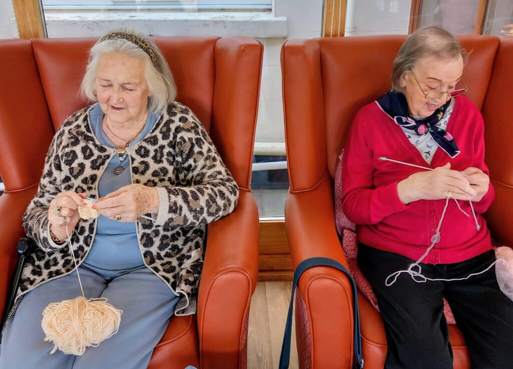 Two elderly women sit in orange armchairs, smiling at one another as they concentrate on knitting pale-coloured wool—each stitch adding to heartfelt Shoebox Surprises.
