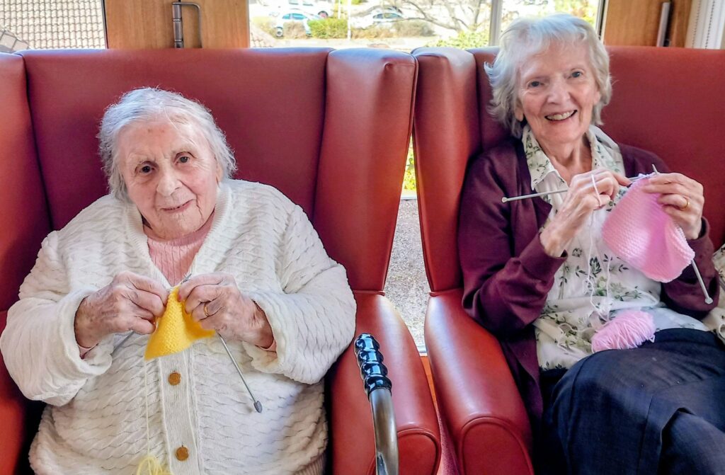 Two elderly women sit side by side in orange chairs, knitting cheerful stitches with yellow and pink wool. Both are smiling and seem to be in a bright indoor setting.