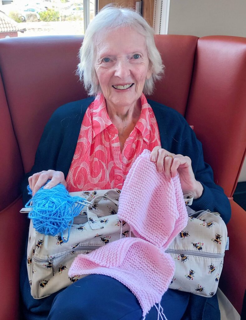 An elderly woman sits in a red armchair, smiling and holding pink knitting and blue wool on her lap as she adds careful stitches for Shoebox Surprises, with a patterned bag next to her.