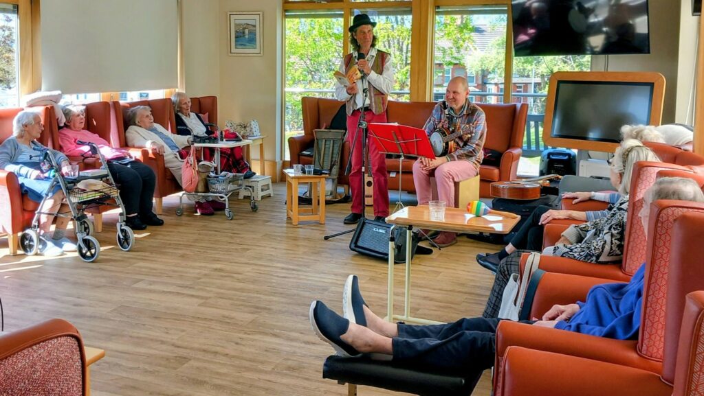 Two musicians perform for a group of elderly people seated in a brightly lit lounge with large windows and comfy chairs.