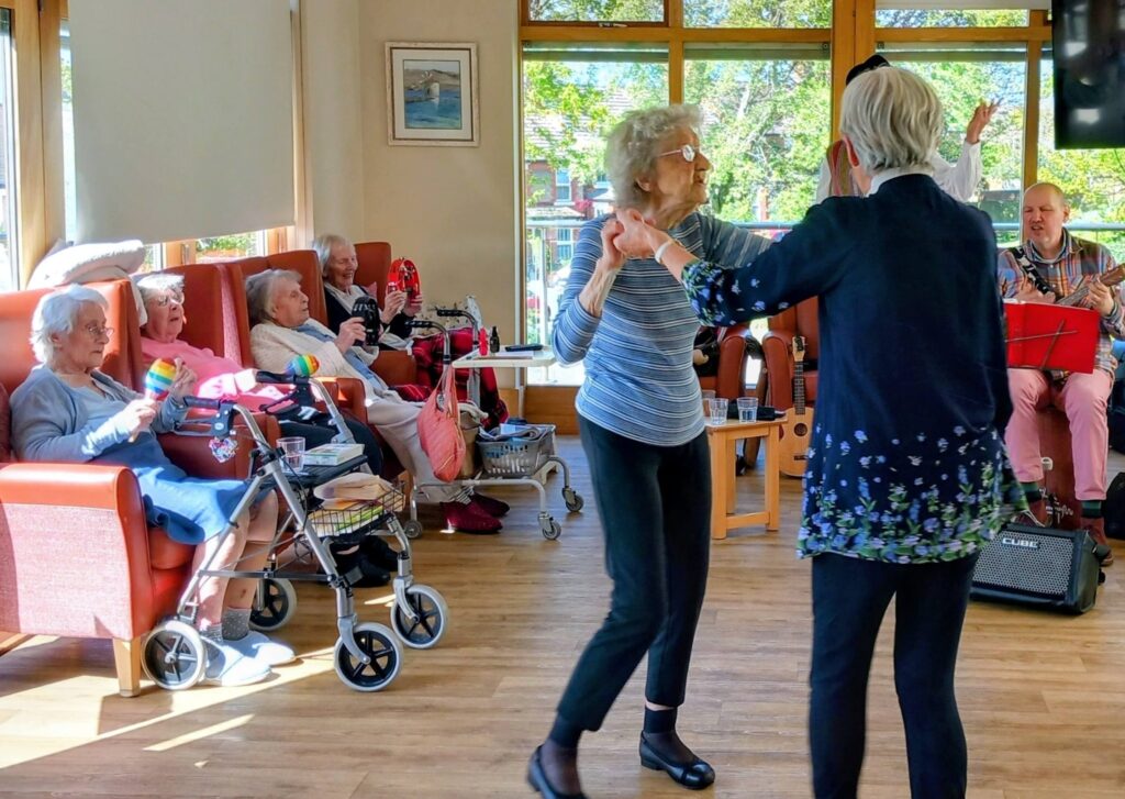 Two elderly women dance together in a sunlit room whilst other pensioners sit and watch; a small musical group performs with instruments in the background.