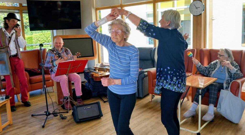 Two elderly women dance together in a bright room while two musicians perform; another woman sits nearby watching and holding a maraca.