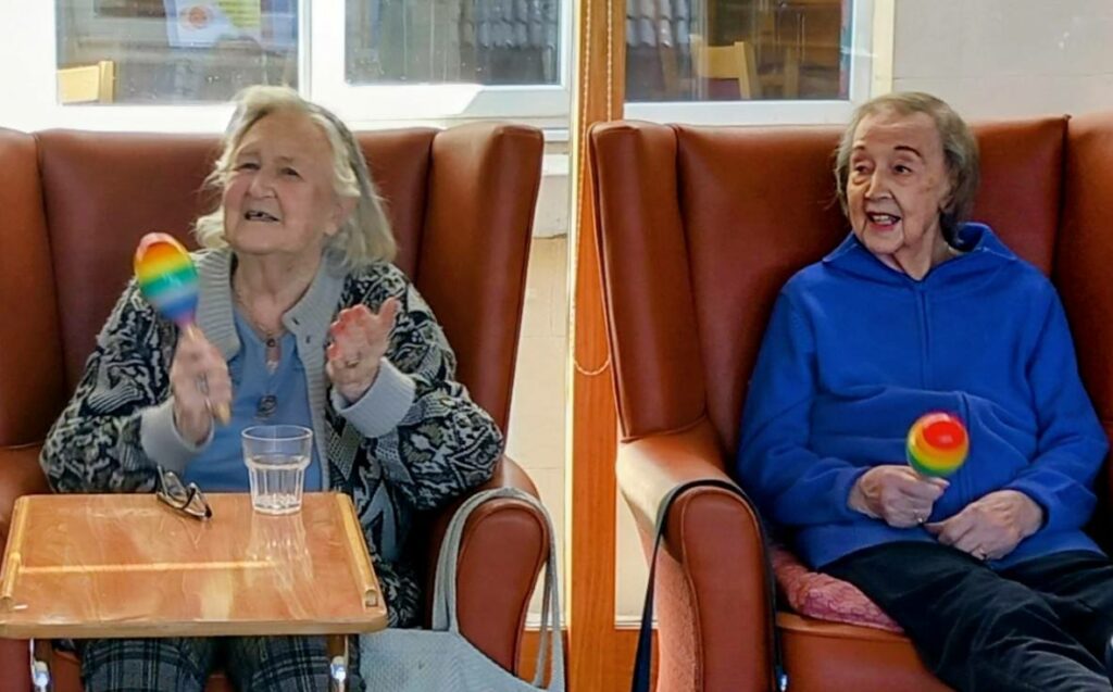 Two elderly women sit in armchairs, smiling and holding rainbow-coloured maracas, with a table and a glass of water in front of one of them.