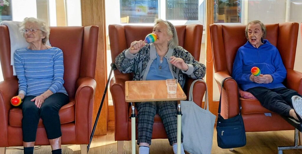 Three elderly women are seated in armchairs; two of them hold colourful maracas and seem to be taking part in a group activity.