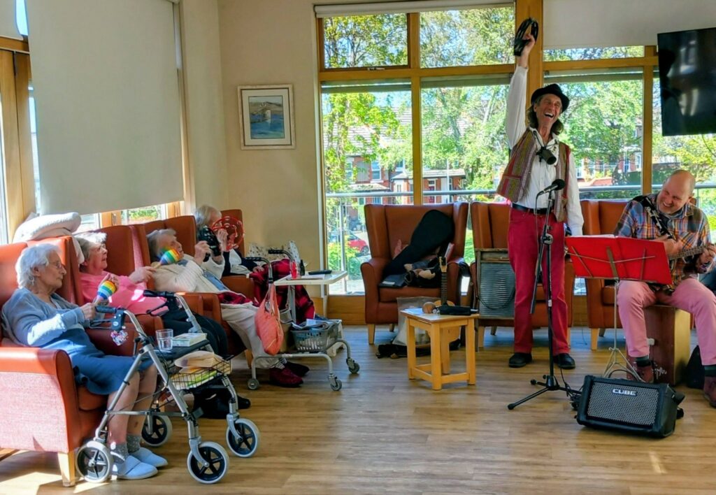 Entertainers perform for a group of elderly people in a bright room; one stands singing with a tambourine whilst the other plays guitar. Pensioners sit in armchairs, some with walking frames.