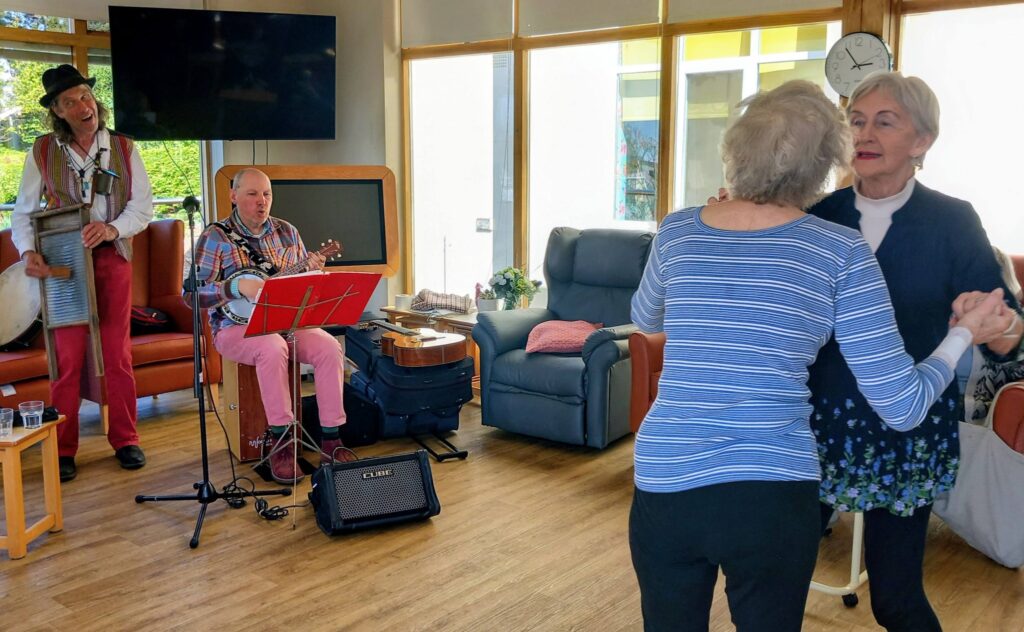 Two elderly women dance together in a well-lit room whilst two musicians perform, one playing a washboard and the other a banjo.