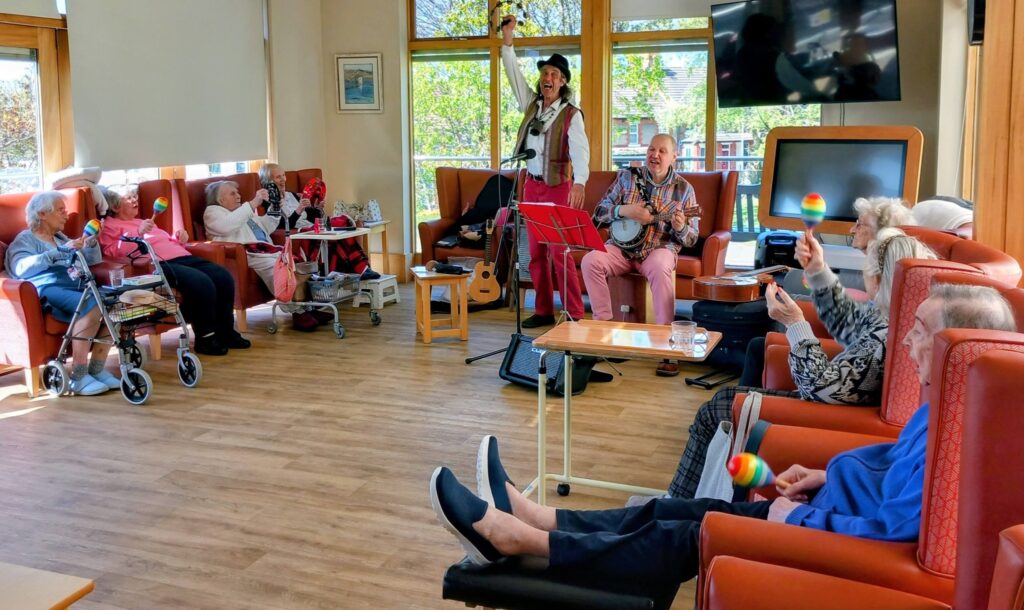 Elderly people sit in a bright communal room watching two musicians perform; some hold maracas and clap whilst others relax in armchairs.