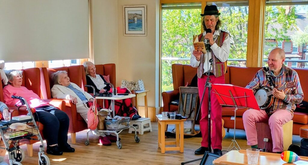 Two musicians entertain four seated elderly people in a bright room; one plays the banjo while the other speaks into a microphone and holds a book.