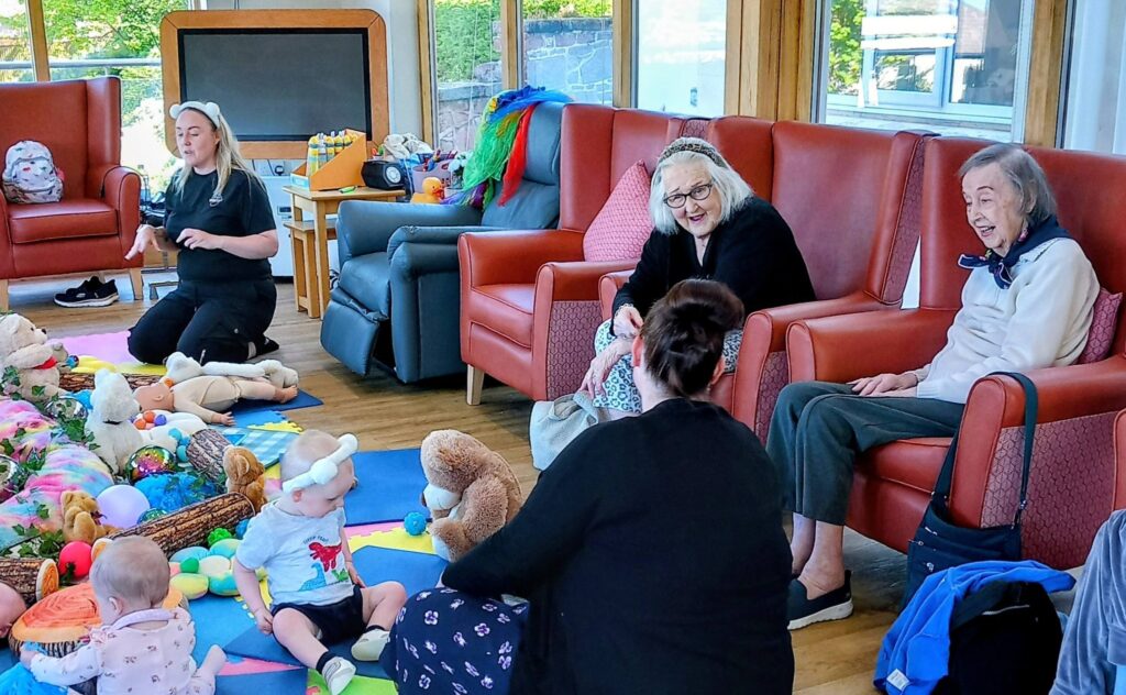 Older adults and carers sit on armchairs, engaging with babies and young children who are playing with toys on mats in a brightly lit room.