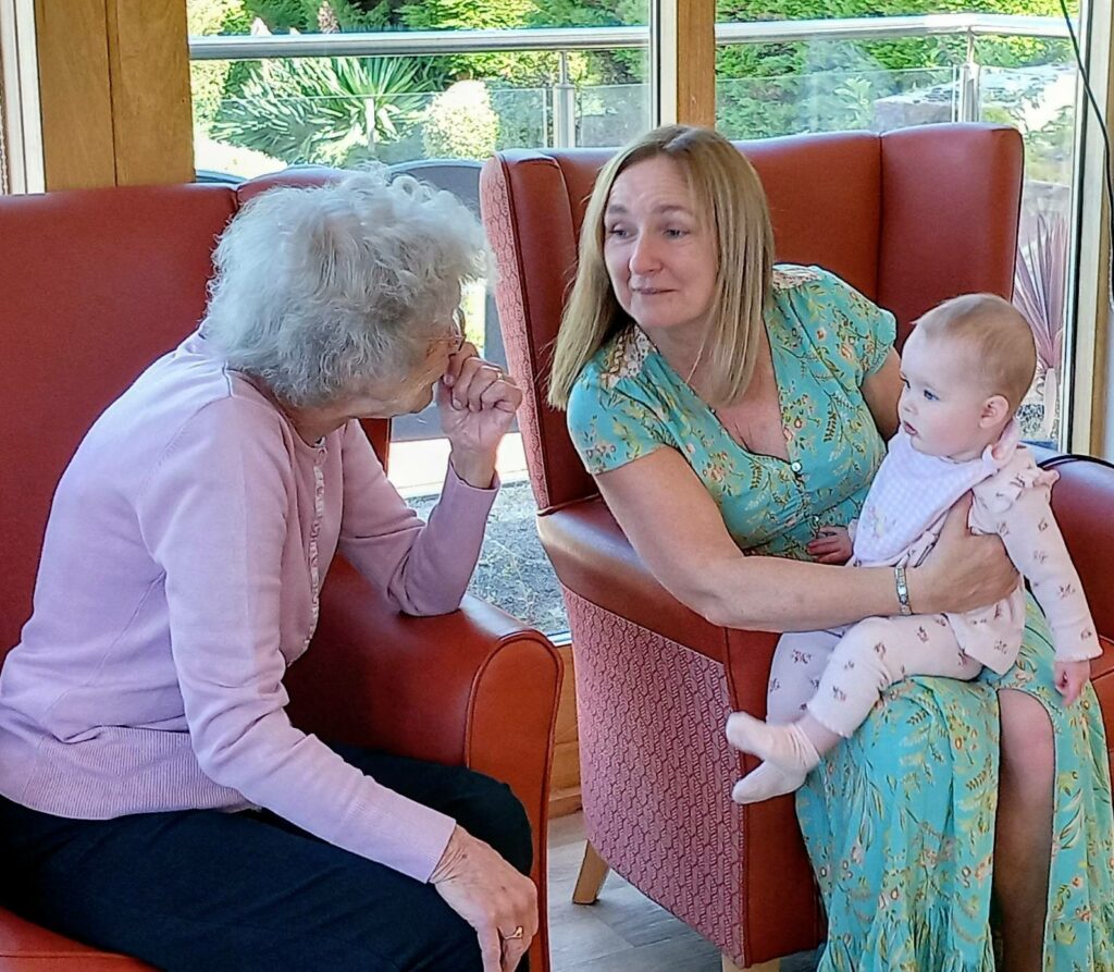 An elderly woman sits in a red armchair, chatting to a woman who has a baby on her lap, in a bright room with large windows.