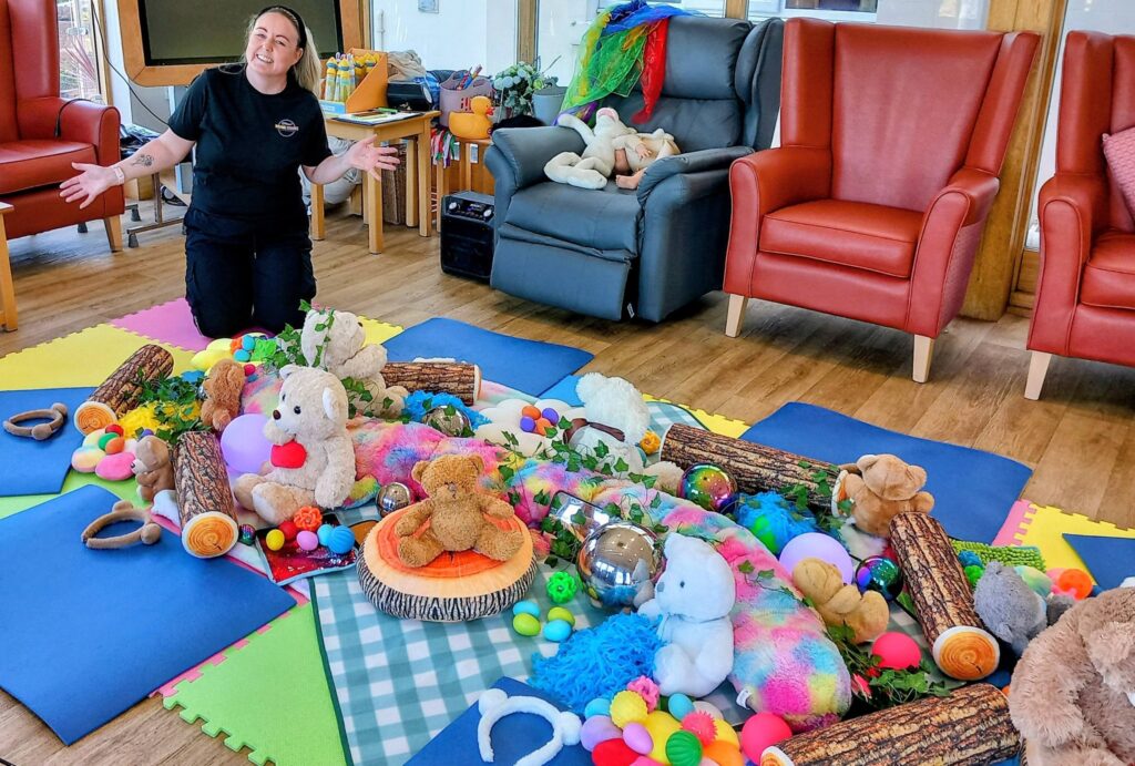 A woman kneels on colourful foam mats surrounded by soft toys, balls and sensory playthings in a cosy room with red chairs and gentle lighting.