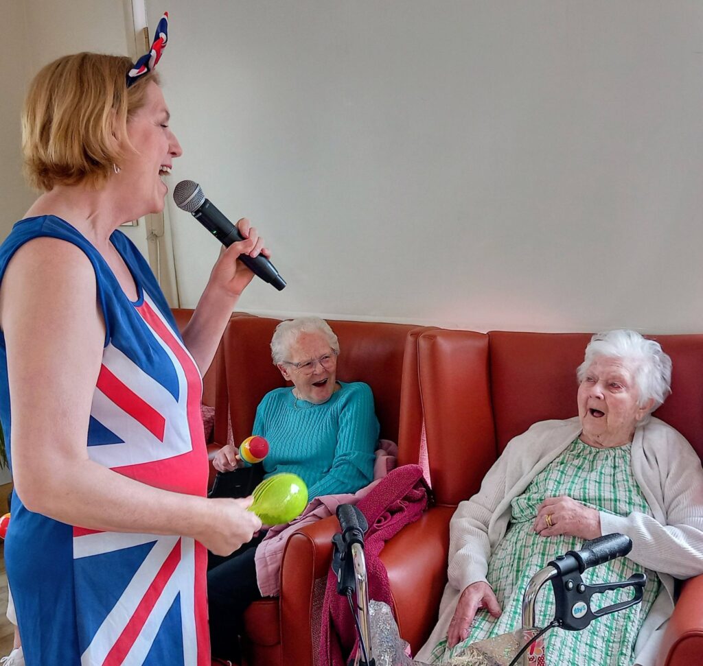 A woman in a Union Jack dress sings into a microphone at Lear House, entertaining two elderly ladies, including Thelma, who seem to be enjoying the birthday celebration.