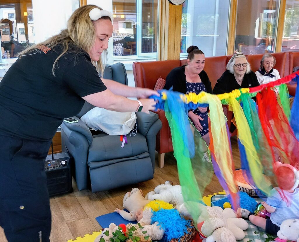 A woman waves colourful scarves in front of seated adults and a baby on a play mat with soft toys, all in a bright, communal room.
