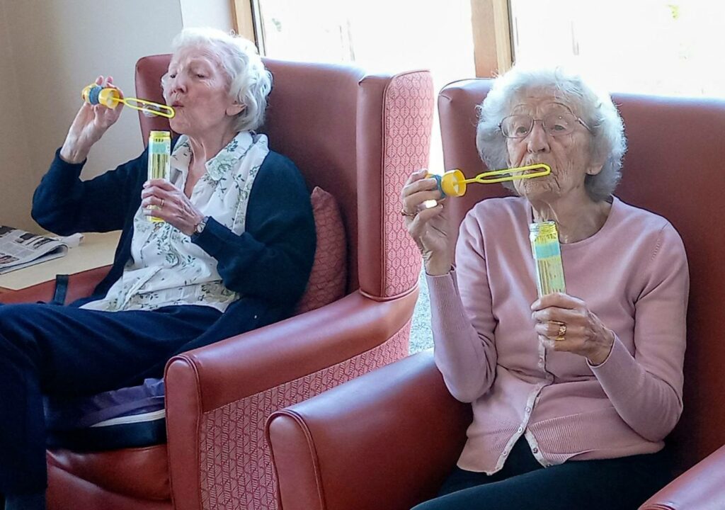 Two elderly women seated in armchairs blow bubbles with bubble wands.