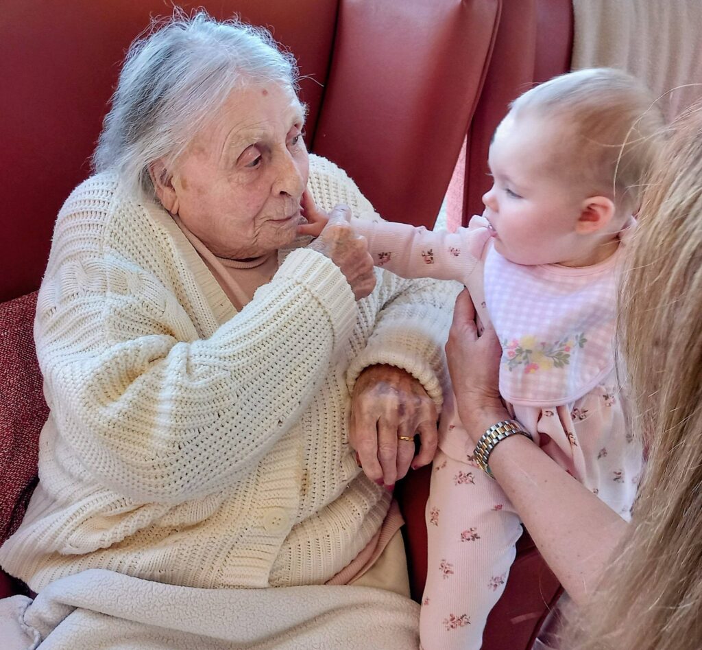 An elderly woman in a cream jumper sits in a chair, gently touching the hand of a baby dressed in pink, who is being held by an adult.
