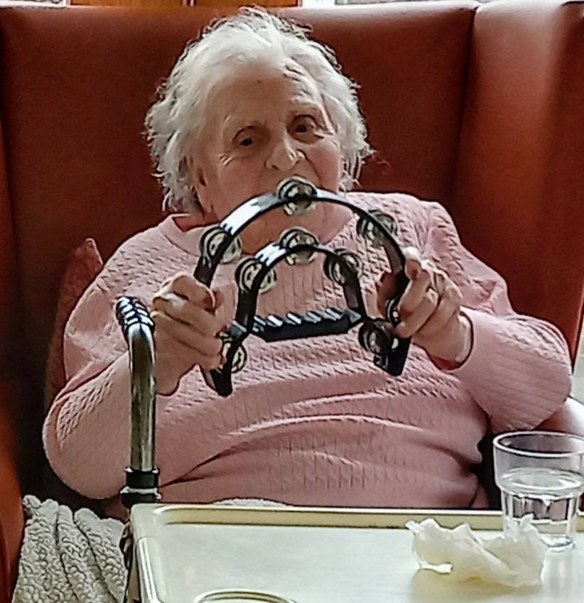 Thelma, at Lear House, sits in her armchair wearing a pink jumper and holding a tambourine. A walking frame, glass of water, and tissues are on the table in front of her during her birthday celebration.