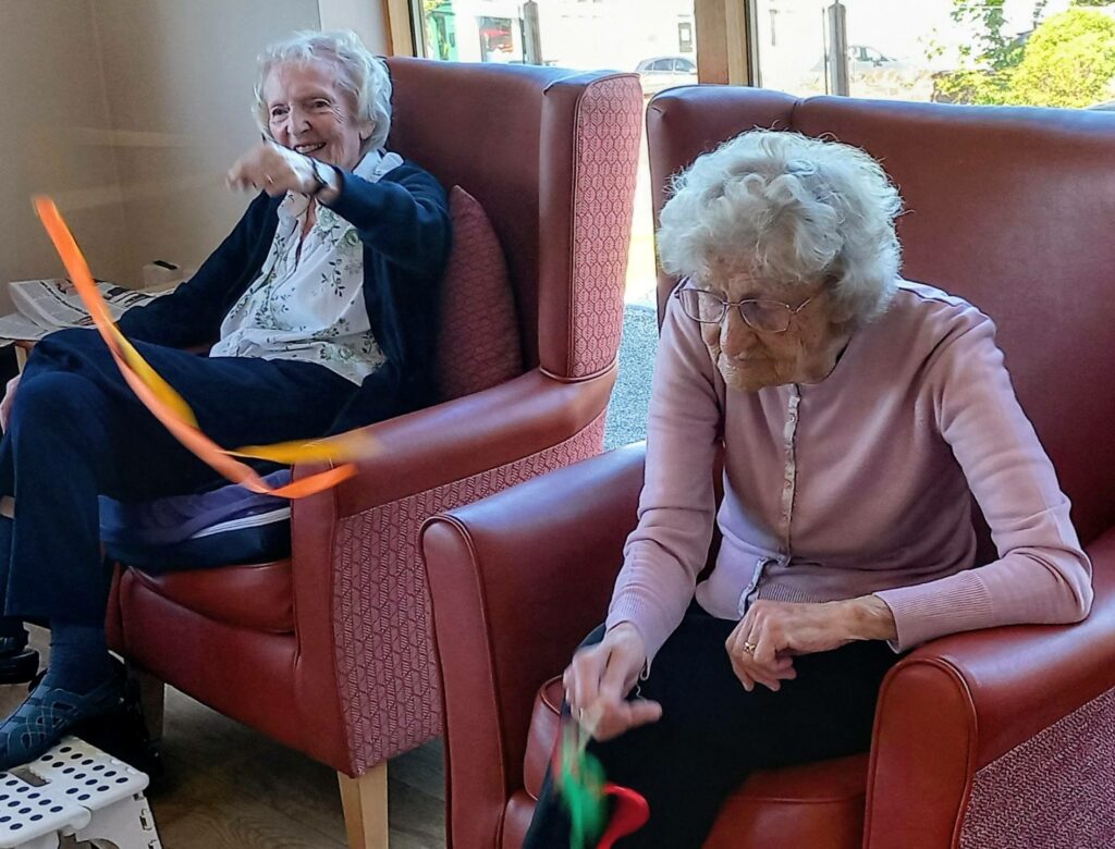 Two elderly women seated in armchairs wave colourful ribbons, taking part in an activity in a well-lit room.