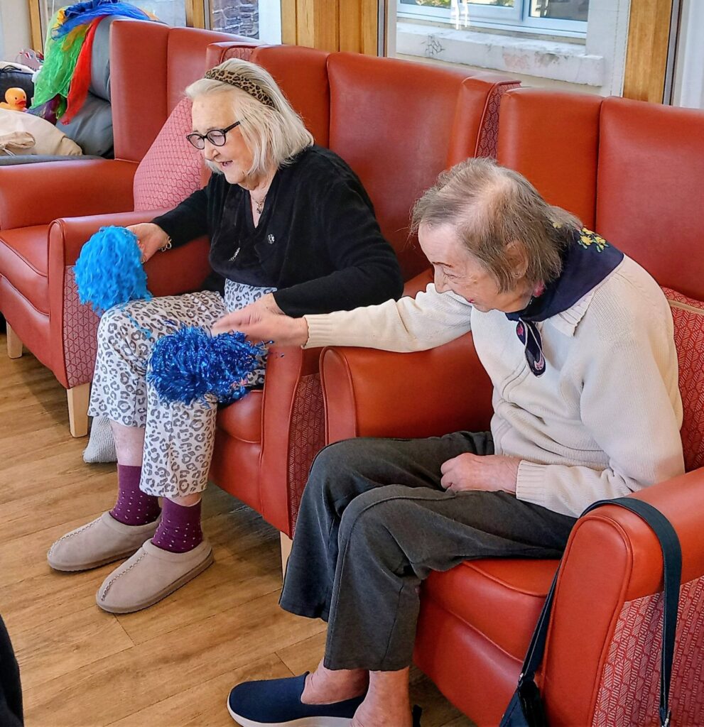 Two elderly women sit on red chairs indoors, holding blue pom-poms and engaging with one another.