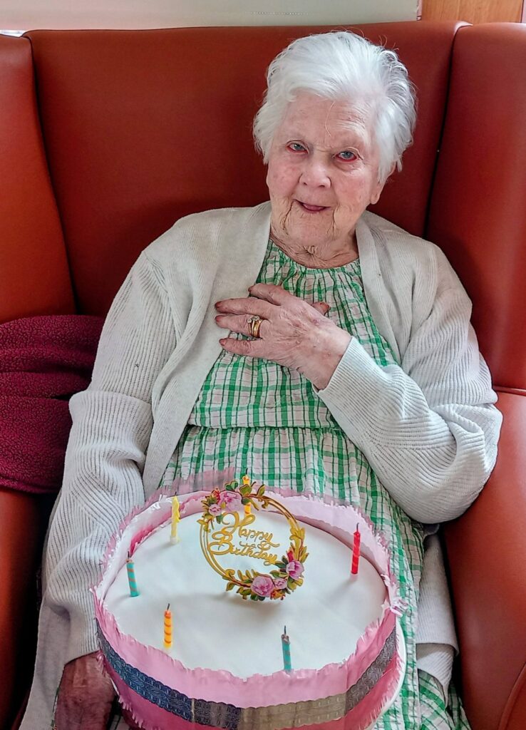 Thelma, an elderly woman at Lear House, sits on an orange chair wearing a checked dress and cardigan, holding a birthday cake with candles and a “Happy Birthday” topper for her birthday celebration.