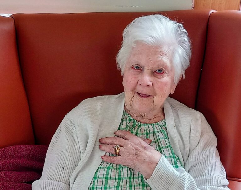Thelma, an elderly woman at Lear House, sits on an orange chair wearing a checked dress and cardigan, holding a birthday cake with candles and a “Happy Birthday” topper for her birthday celebration.