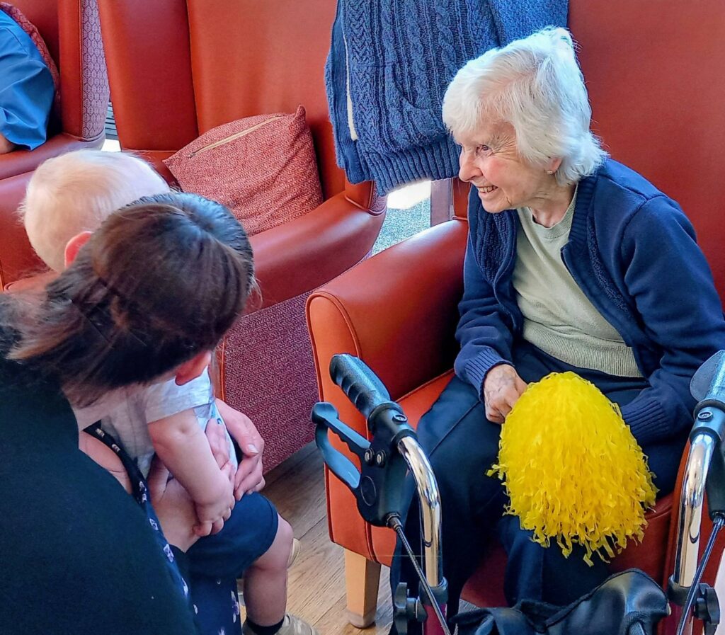 An elderly woman holding a yellow pom-pom sits in an armchair, smiling at a baby being held by a woman with her back to the camera. A walking frame is positioned next to the elderly woman.
