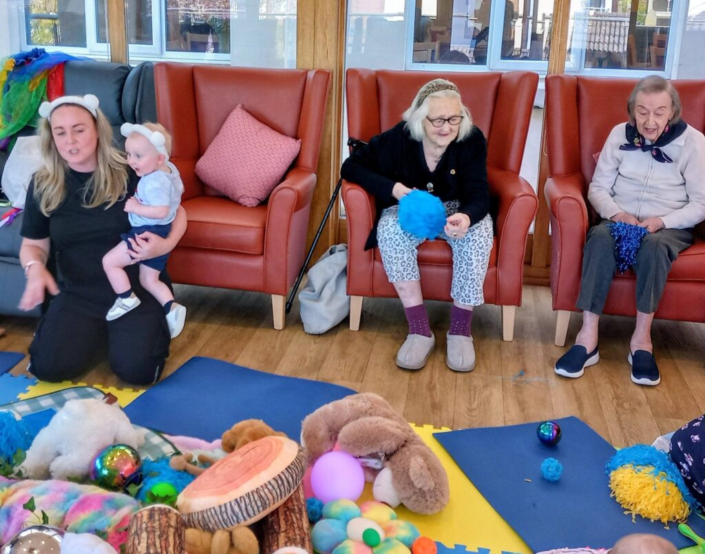Three adults and a baby are seated in a lounge with red chairs, surrounded by colourful toys, balls, and soft toys on foam mats.
