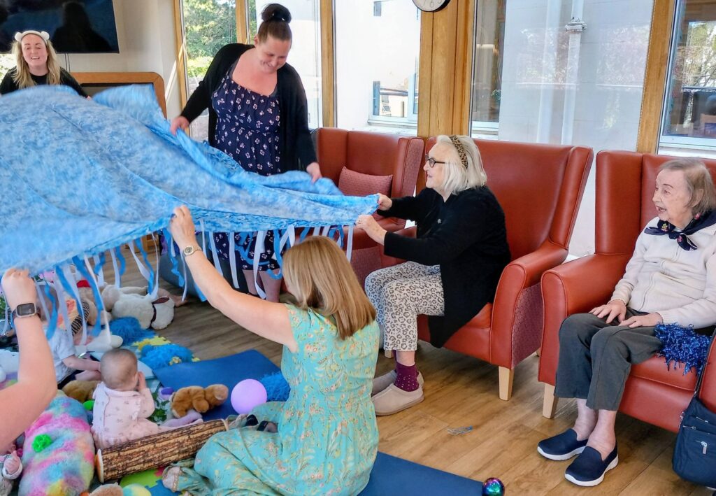 Several adults and babies sit and play on the floor with soft toys, whilst two women hold up a blue fabric sheet. Two elderly adults sit watching from red armchairs.