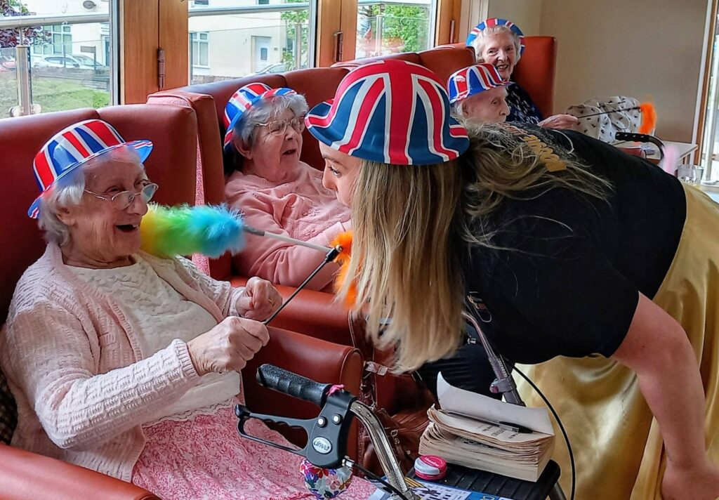 A group of elderly women sporting hats adorned with the UK flag share a laugh at Lear House, as one playfully brushes another’s face with a colourful feather duster, embodying the Best of British spirit.