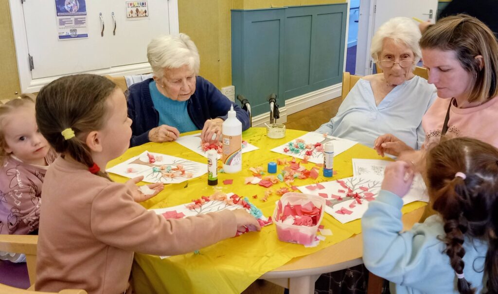 Two elderly women and three young visitors sit around a table enjoying a colourful morning of arts and crafts with paper, glue, and colouring sheets.