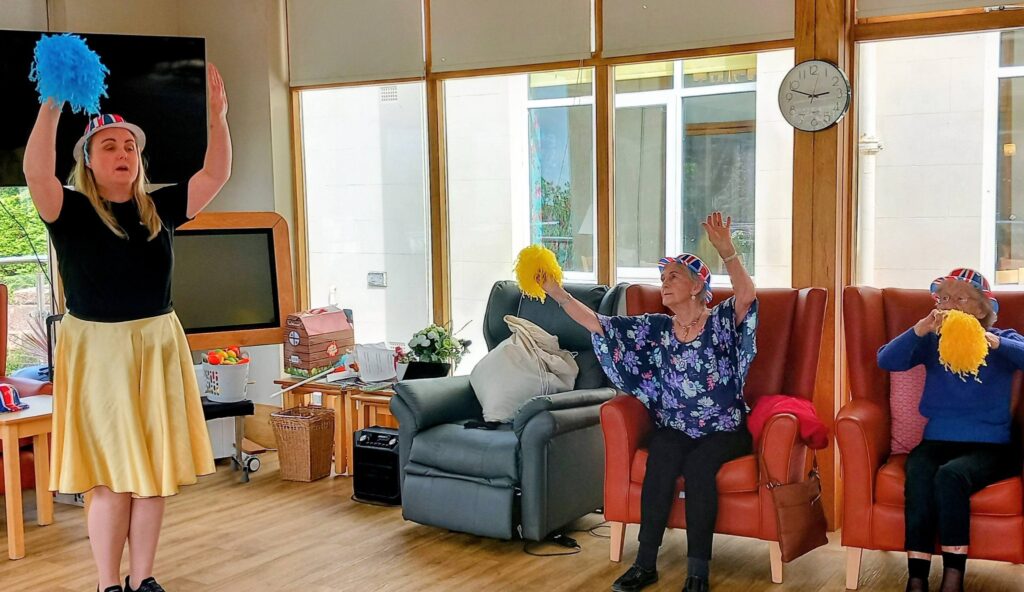 At Lear House, a woman leads two elderly people in seated exercises with pom-poms. All three are wearing festive hats and have their arms raised, celebrating the Best of British spirit.