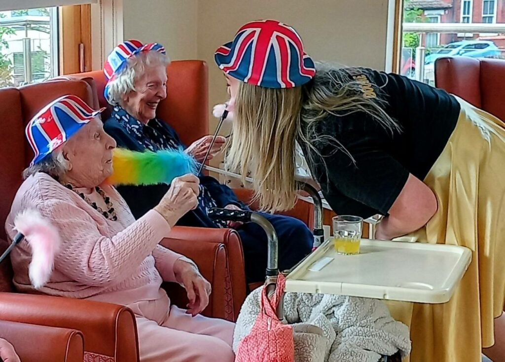 Two elderly women at Lear House, donning Union Jack hats, sit in armchairs as one playfully waves a colourful feather—perfectly capturing the cheerful spirit of Best of British.